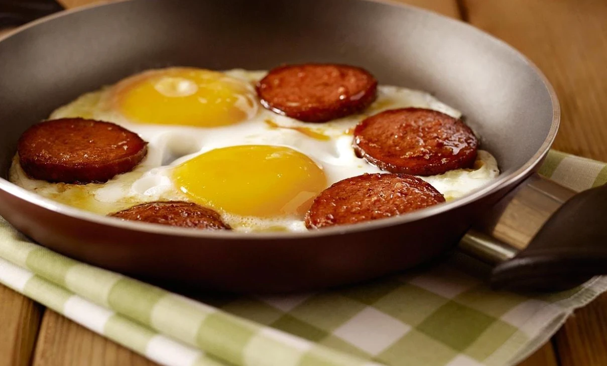 Oval sahanda pişmiş yumurta ve kızarmış sucuk dilimleri, yanında ekmek ve çeri domatesler